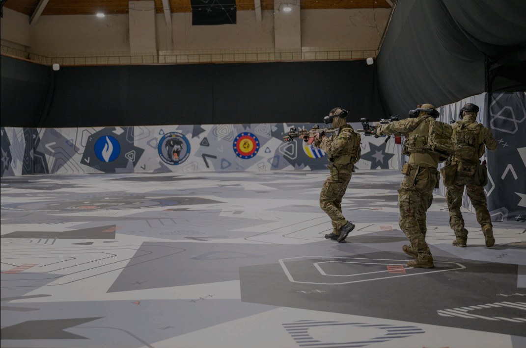 Three people in camouflage wearing VR headsets and holding rifle-shaped controllers, in a hall with a marked floor